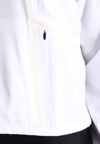 Zbliżenie na białą kurtkę, pokazujące pionową kieszeń z przodu zapinaną na suwak z czarnym uchwytem oraz delikatny beżowy pionowy pasek.