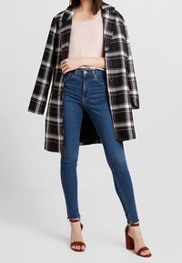 Svart och vit rutig kappa med röda detaljer, kombinerad med en ljusrosa topp och blå skinny jeans. Har blockklackade bruna sandaler.