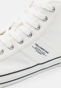 Biały, wysokoprofilowy trampek z kontrastową niebieską podeszwą, okrągłymi metalowymi oczkami i oznaczoną boczną metką z napisem „YELLOWSTONE” oraz współrzędnymi.