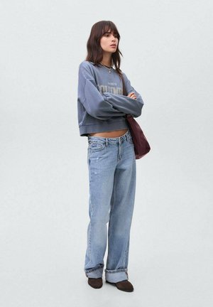 Felpa grigia cropped con testo, jeans a palazzo blu chiaro e scarpe slip-on marroni. Modello in posa con le braccia incrociate, porta una borsa bordeaux.
