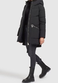 Manteau noir en duvet avec capuche, doté de zip argentés et de poches latérales. Porté avec un jean skinny noir et des bottines noires à semelle épaisse.