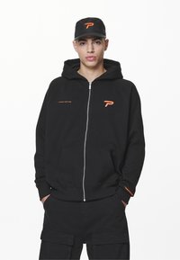 Svart zip-up hettegenser med oransje logo på brystet, framlommer og justerbar hette. Kombinert med matchende svarte cargo-bukser og caps.