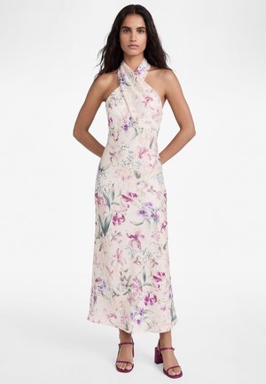 Frau trägt ein ärmelloses, neckholderlanges Kleid mit pastellfarbenem Blumenmuster und violetten High-Heel-Sandalen, steht mit den Händen hinten.