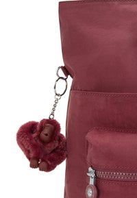 Pluszowy bordowy breloczek w kształcie goryla przymocowany do bocznej kieszeni bordowego plecaka z zamkiem błyskawicznym.