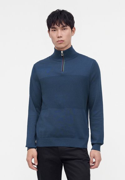 Jeune homme portant un pull en maille bleu foncé avec un panneau texturé sur la poitrine et un col à demi-fermeture éclair, associé à un pantalon noir, debout sur un fond neutre.