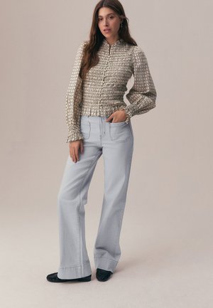 Femme portant un chemisier à manches longues à motifs avec une taille smockée et un pantalon large bleu clair, debout avec une main dans la poche.