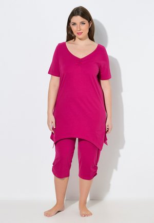 Femme debout pieds nus portant une tunique à manches courtes magenta et des leggings raccourcis assortis, sur un fond blanc uni.