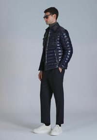 Doudoune marine avec texture matelassée, poches avant et boutons-pression, associée à un pantalon noir et des baskets blanches. Modèle portant des lunettes de soleil.