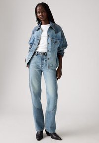 Jachetă și blugi din denim albastru, ambele cu un aspect decolorat. Tricou alb sub jachetă. Curea neagră și cizme cu toc ascuțit. Fundal simplu.