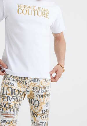 White short-sleeve shirt with gold "Versace Jeans Couture" text paired with white patterned pants featuring gold baroque designs and black text.