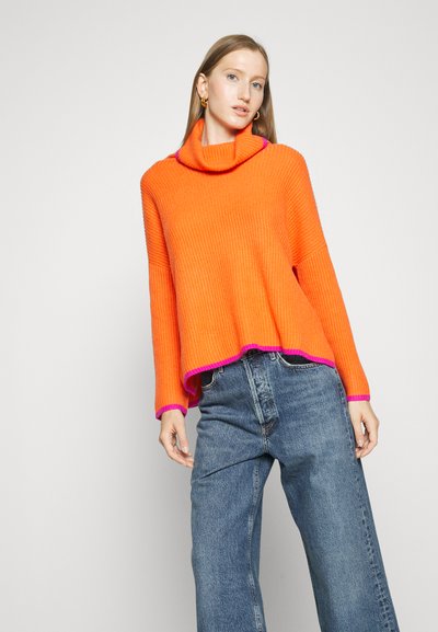 Orange ribbed sweater featuring a cowl neck and bright pink accents at the cuffs and hem, paired with loose-fitting blue jeans.