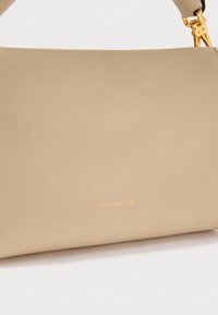 Smėlio spalvos tekstūruota odinė rankinė su auksinėmis detalėmis ir „Coccinelle“ išgraviruotu mažu auksiniu užrašu priekyje.