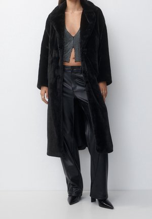 Femme portant un long manteau en fausse fourrure noir sur un pantalon en cuir noir, un top court noir et blanc, et des talons pointus noirs.