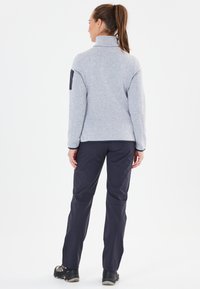 Graue Fleecejacke mit hohem Kragen und schwarzen Akzenten an den Ärmeln, kombiniert mit dunkler Hose und robustem schwarzen Schuhwerk.