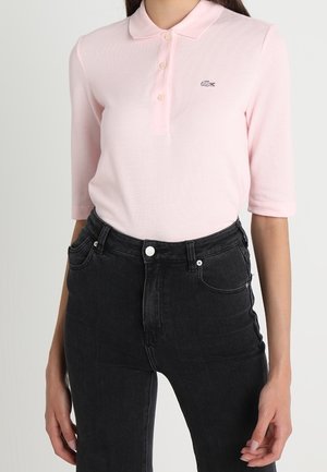 Hellrosa Poloshirt mit einer dreiknöpfigen Knopfleiste und einem auf der Brust gestickten Logo, kombiniert mit hoch taillierten schwarzen Jeans aus Denim.