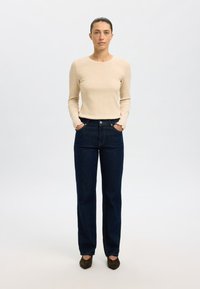 Bézs színű hosszú ujjú bordázott felső, sötétkék magasderekú egyenes szárú farmerrel párosítva. A cipők sötétbarnák, lekerekített orrú kialakítással.