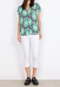 Blusa a maniche corte verde e grigia con motivo, colletto rotondo, abbinata a pantaloni bianchi cropped e scarpe nere con accenti dorati.
