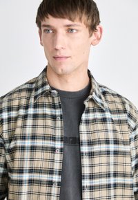 Beige plaid shirt with blue and black checks, button-down collar, and multicolored buttons, layered over a gray t-shirt with printed logo.