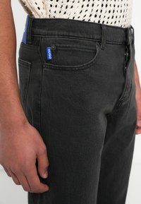 Mustad denimist kitsas lõikega teksad, millel on vöökohal sinine kaubamärgiga silt ja esitaskutel peened õmblused.