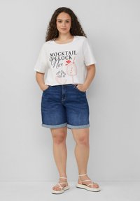 Witte graphic T-shirt met de tekst "MOCKTAIL O'CLOCK" en een cocktailontwerp, gecombineerd met donkerdenim shorts met opgerolde zoom en witte strapsandalen.