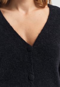 Zwarte wollen cardigan met een V-halsontwerp, zachte textuur en een enkele knoopsluiting aan de voorkant. Close-up weergave van de stofdetails.