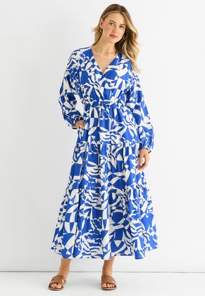 Maxi-jurk in blauw-witte patronen, met lange mouwen, een getailleerde taille, een gelaagde rok en lichtgewicht stof. Lichtbruine sandalen.