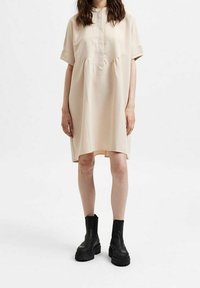 Béžové šaty s krátkým rukávem, knoflíkovou légou, nařaseným pasem a postranními rozparky, v kombinaci s robustními černými kotníkovými botami. Hladká textilie.