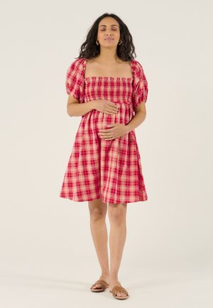 Schwangere Frau in einem roten karierten Kleid mit Puffärmeln hält sanft ihren Bauch und steht barfuß vor einem schlichten Hintergrund.