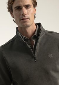 Sudadera de cremallera de color verde oscuro con tejido texturizado, cuello alto y logo en el pecho. Llevada sobre una camisa de botones color gris claro.