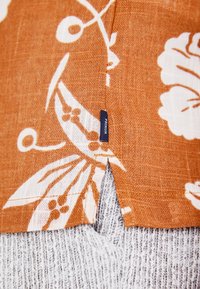 Tela de lino naranja con patrones florales blancos, que presenta una abertura lateral y una pequeña etiqueta azul en el dobladillo. Tela texturizada gris debajo.