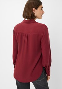 Burgundy long-sleeve blouse with a collared neckline, pleated back detail, and rounded hem, made of soft fabric with a smooth texture.