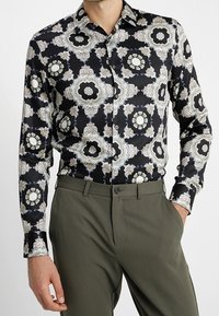 Chemise noire à boutons avec un motif composé de formes florales complexes en blanc, gris et bleu. Associée à un pantalon vert olive.