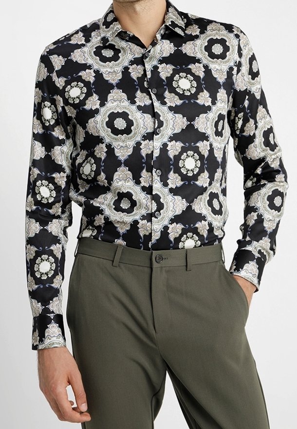 Chemise noire à boutons avec un motif composé de formes florales complexes en blanc, gris et bleu. Associée à un pantalon vert olive.
