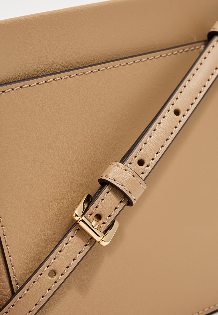 Beige leren tasdetail met een gestructureerde afwerking, voorzien van een gouden gesp en band. Zichtbaar stikwerk voegt ontwerpelementen toe.