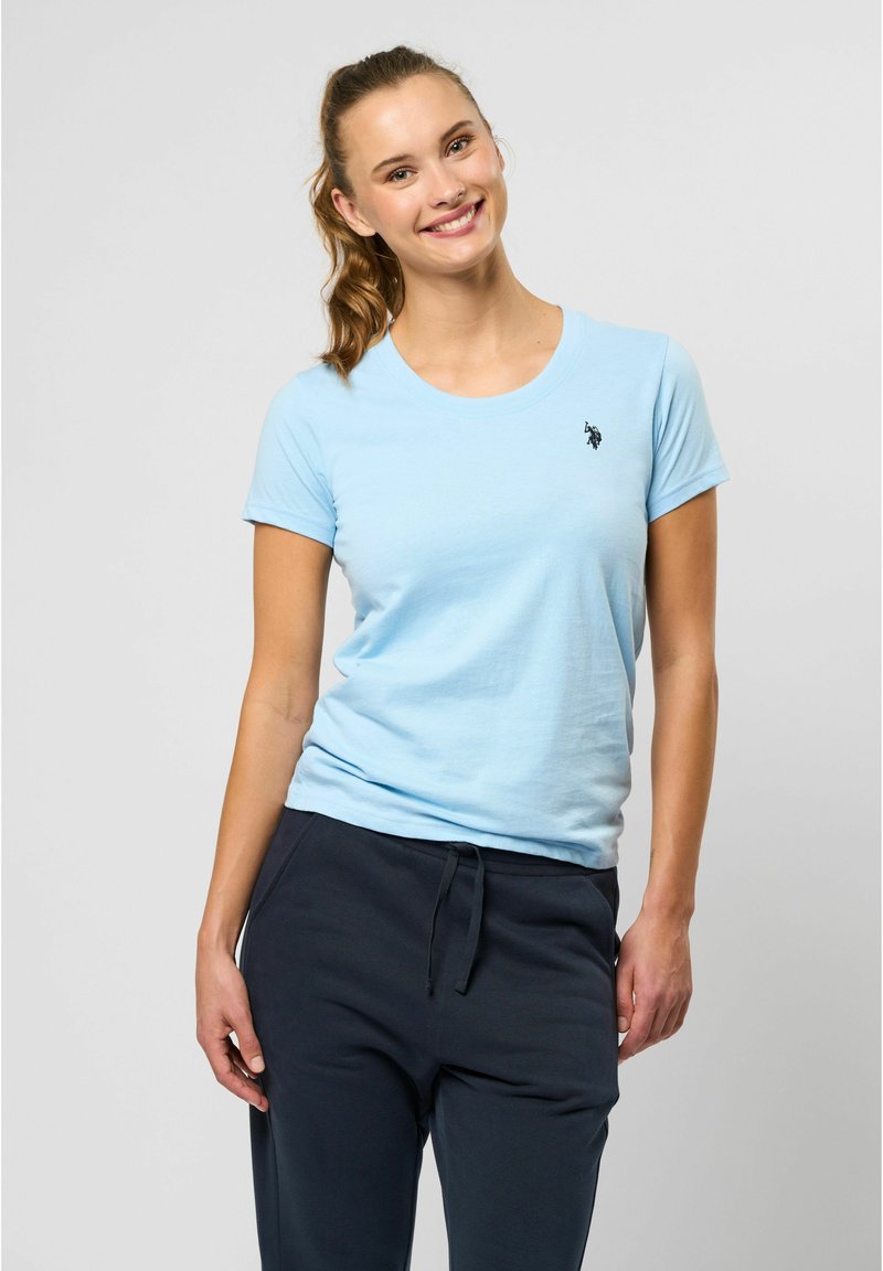 Lyseblå bomulls T-skjorte med korte ermer og rund hals, med en liten svart logo på brystet. Kombinert med mørke joggebukser.