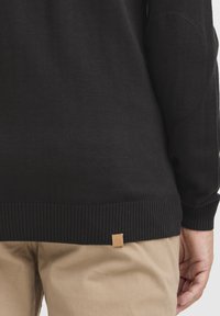 Svart stickad tröja med ribbade ärmslut och nederkant, med en läderetikett i detalj vid botten, samt armbågslappar i ett liknande tyg.