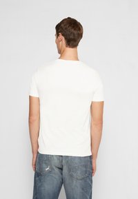 Bílé bavlněné tričko s krátkými rukávy a kulatým výstřihem, zobrazené zezadu. Nošeno s modrými džínami s bočními kapsami vzadu.