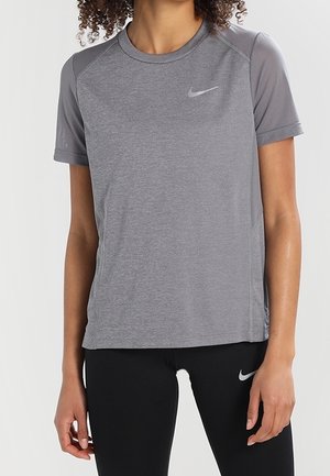 Frau trägt ein eng anliegendes graues kurzärmeliges Sportshirt und schwarze Leggings, beide mit kleinen weißen Markenlogos.
