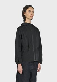 Giacca leggera nera con cappuccio, chiusura frontale con zip, cordini regolabili in vita e polsini elasticizzati. Indossata sopra pantaloni in denim scuro.