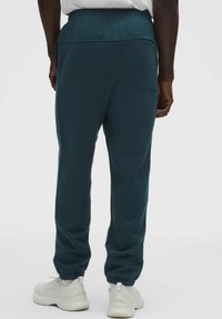 Joggers en verde azulado de tejido suave con cintura elástica y puños ajustables, que cuentan con un bolsillo trasero y un corte holgado. El calzado es de color blanco.