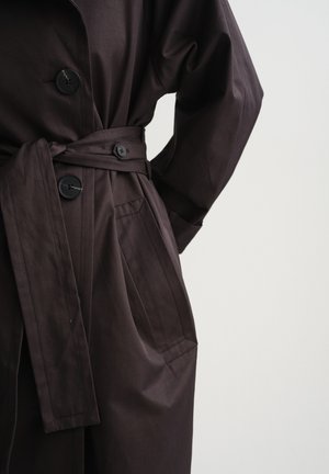 Dark brown buttoned trench coat with waist belt and side pocket, worn with hand resting on hip against plain background.