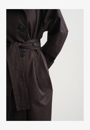 Dark brown buttoned trench coat with waist belt and side pocket, worn with hand resting on hip against plain background.