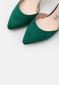 Zelené semišové lodičky s špičatou špičkou, hladkým povrchem a kovovými akcenty na podpatku. Interiér je v světlé, neutrální barvě.
