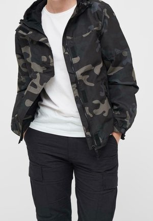 Person iført en sort og grå camouflagejakke over en hvid t-shirt og sorte cargobukser med hænderne i lommerne.