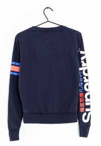Sudadera azul marino con dobladillo y puños acanalados, que presenta un parche rectangular en la parte posterior y un texto en blanco y rojo de gran tamaño a lo largo de la manga.