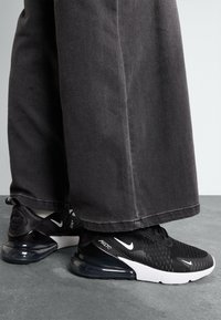 Black Nike Air Max 270 sneakers with a textured upper, white accents, and a transparent air cushioning unit, paired with dark wide-leg trousers.