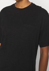 T-shirt noir à manches courtes en tissu doux, avec une petite poche sur la poitrine et un col rond classique. Texture lisse.