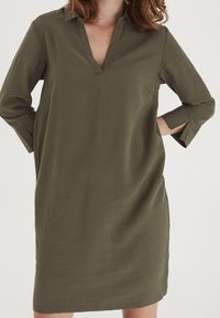 Robe olive verte, longueur genoux avec un décolleté en V et un col. Présente des manches trois-quarts avec des fentes, fabriquée dans un tissu lisse et léger.