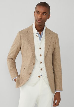 Blazer texturizado marrón claro sobre un chaleco blanco acolchado con detalles de botones, combinado con una camisa a rayas azul y blanca, y pantalones blancos.