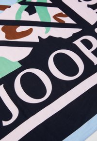 Többszínű textília merész geometrikus mintával, zöld, barna és sötétkék elemekkel, nagy fehér felirattal sötét háttéren.